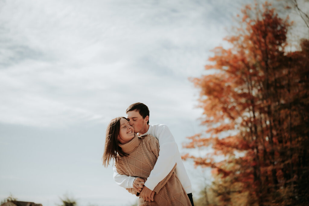 couple pose: a man hugs a woman from behind and planting a kiss on her cheeks.,