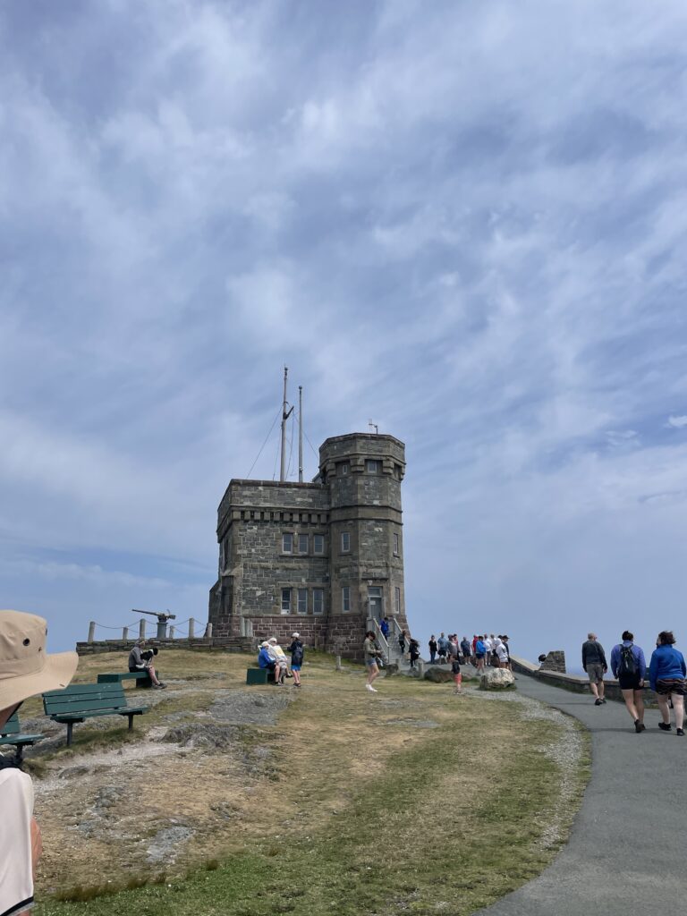 Cabot Tower at the top of Signal Hill on a sunny day, with families taking a break near the wooden stairs.
